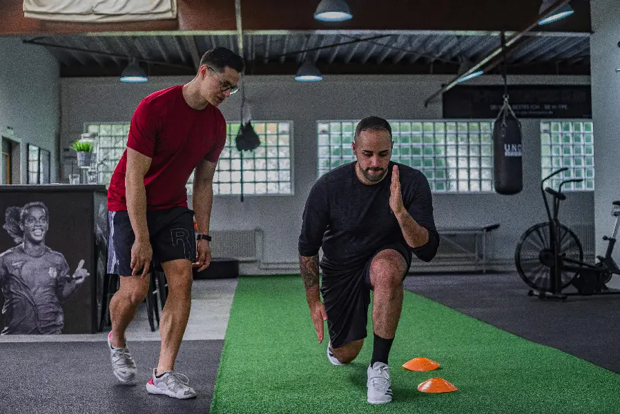 Trainer supervising a person performing a forward lunge drill with cones in a gym training area