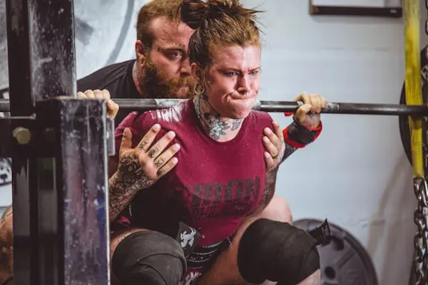Person performing a barbell squat while being assisted by a strength coach in a gym